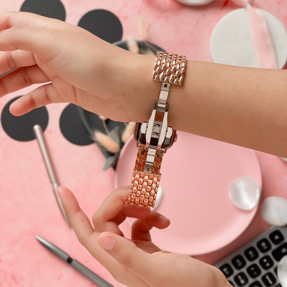 A person is adjusting a Coconut Lane Mermaid Watch Strap in Rose Gold on their left wrist. The background features a pink dish, a calculator, black circular shapes, and a pen. The watchband showcases a woven metallic design and includes a FREE link remover tool. The overall setting seamlessly blends office and personal items.