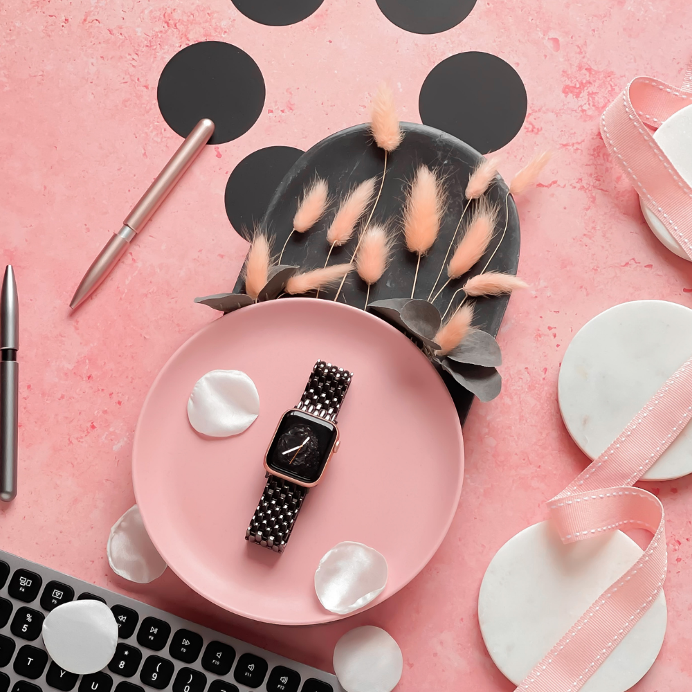 A flat lay on a pink textured surface featuring the Coconut Lane Mermaid Apple Watch Strap in black with a checkered band on a pink plate, surrounded by white petals, a corner of a black keyboard, a silver pen, and decorative dried flowers in a gray vase. Ribbons and marble coasters are also present.