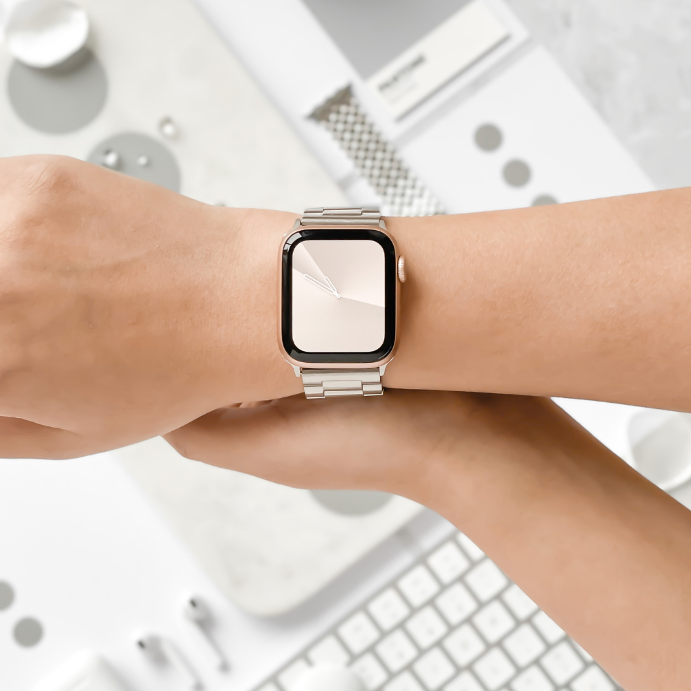 A person is wearing a Coconut Lane Stainless Steel Apple Watch Strap in Silver on their left wrist. The watch features a rectangular face with a minimalistic, sleek design. The background includes blurred office supplies, such as a keyboard, a case, and other gadgets.