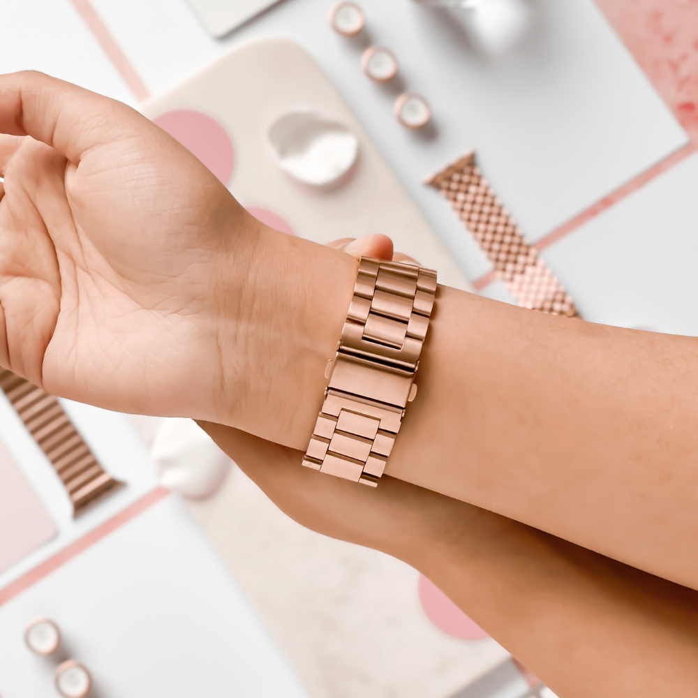 Close-up of a person putting on a Coconut Lane Stainless Steel Apple Watch Strap in Rose Gold. In the background, various accessories and other watch bands, including adjustable stainless steel metal straps for different Apple Watch models, are scattered on a light-colored surface.