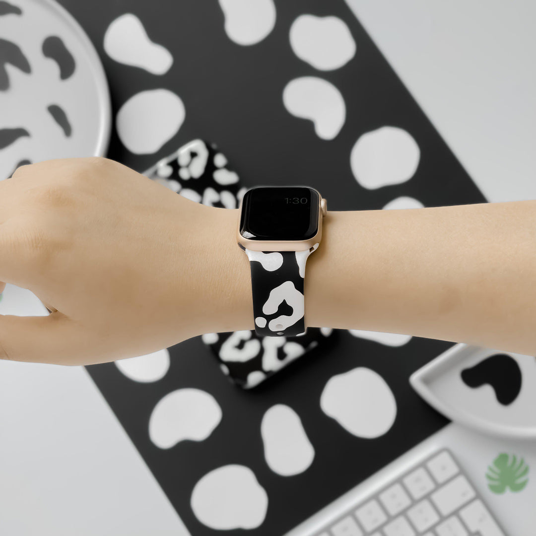 A person wearing a smartwatch with a black and white cow-print band. The background includes items such as a plate, phone case, and keyboard, all featuring the same cow-print pattern. The person is holding their arm horizontally for the photo, showcasing their new Black Leopard Apple Watch Strap from Coconut Lane.