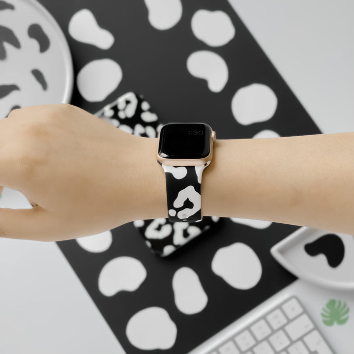 A person wearing a smartwatch with a black and white cow-print band. The background includes items such as a plate, phone case, and keyboard, all featuring the same cow-print pattern. The person is holding their arm horizontally for the photo, showcasing their new Black Leopard Apple Watch Strap from Coconut Lane.