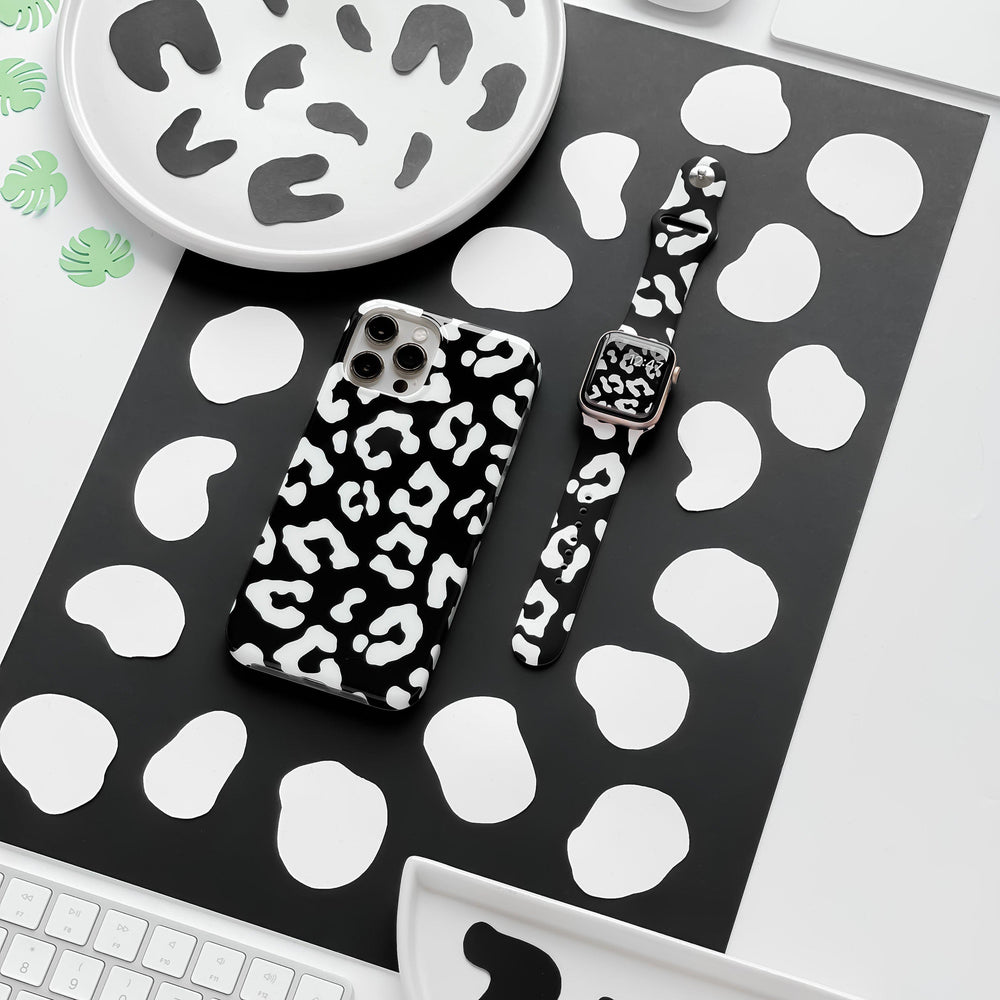 Flat lay of a desk featuring a smartphone with a black and white cow print case and the Black Leopard Apple Watch Strap by Coconut Lane. The desk items rest on a black and white spotted mat, surrounded by plates and coasters with the same black and white pattern.
