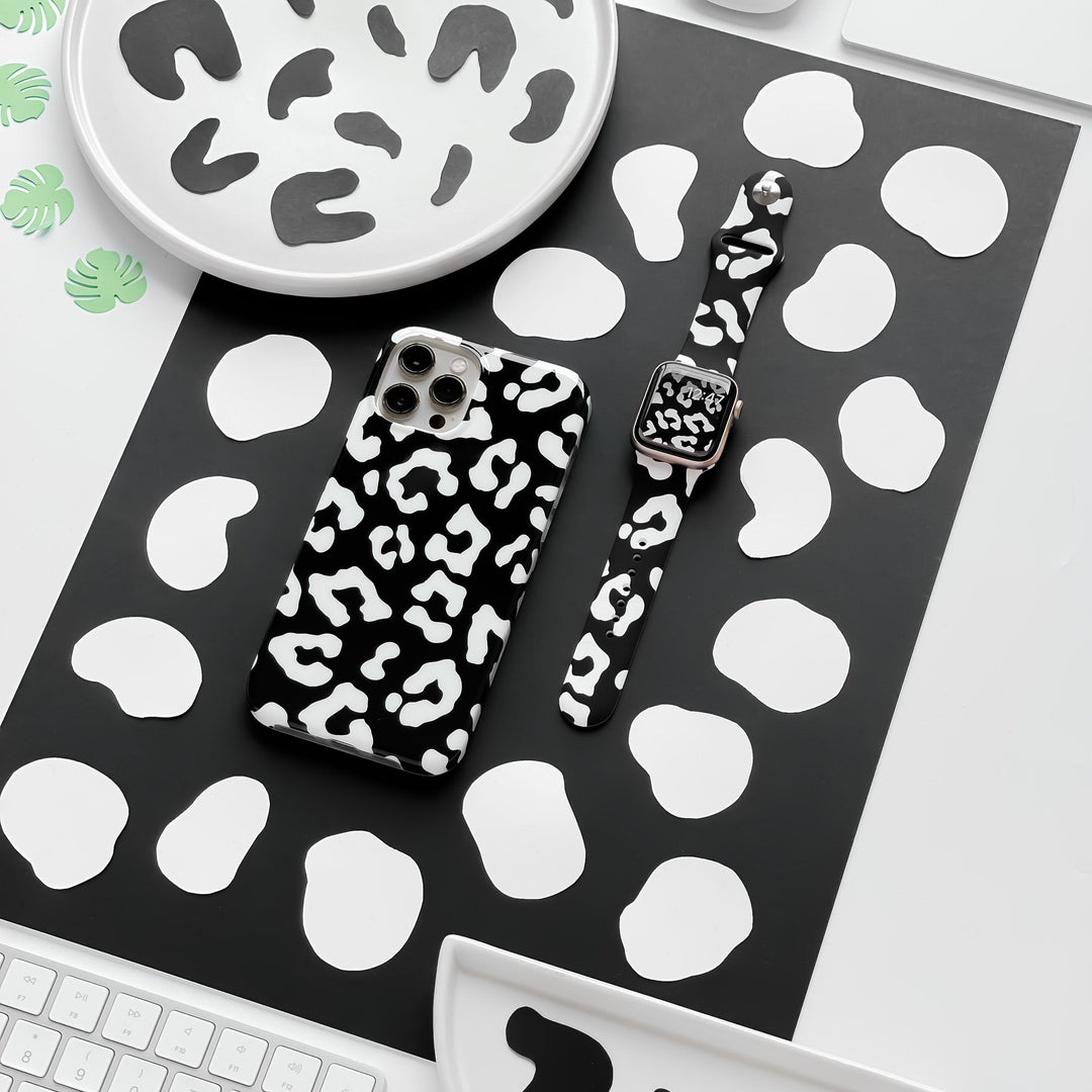 Flat lay of a desk featuring a smartphone with a black and white cow print case and the Black Leopard Apple Watch Strap by Coconut Lane. The desk items rest on a black and white spotted mat, surrounded by plates and coasters with the same black and white pattern.