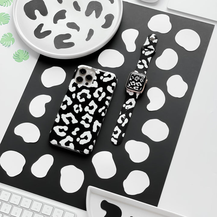 Flat lay of a desk featuring a smartphone with a black and white cow print case and the Black Leopard Apple Watch Strap by Coconut Lane. The desk items rest on a black and white spotted mat, surrounded by plates and coasters with the same black and white pattern.