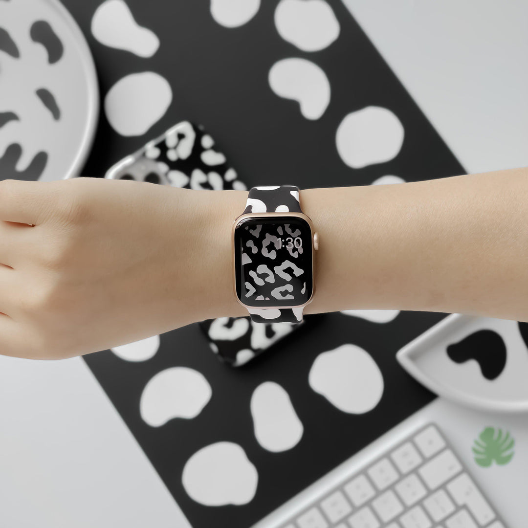 A person's wrist is shown wearing a Coconut Lane Black Leopard Apple Watch Strap, which features a striking black and white abstract pattern. The background includes matching items from the Black Leopard Collection, such as a smartphone case, plates, and a notebook, all showcasing the same bold design alongside a keyboard.