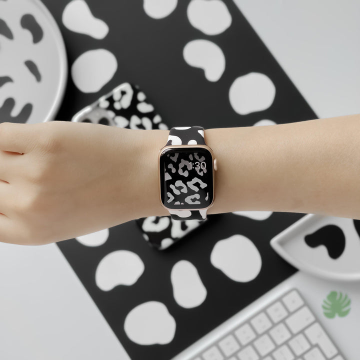 A person's wrist is shown wearing a Coconut Lane Black Leopard Apple Watch Strap, which features a striking black and white abstract pattern. The background includes matching items from the Black Leopard Collection, such as a smartphone case, plates, and a notebook, all showcasing the same bold design alongside a keyboard.