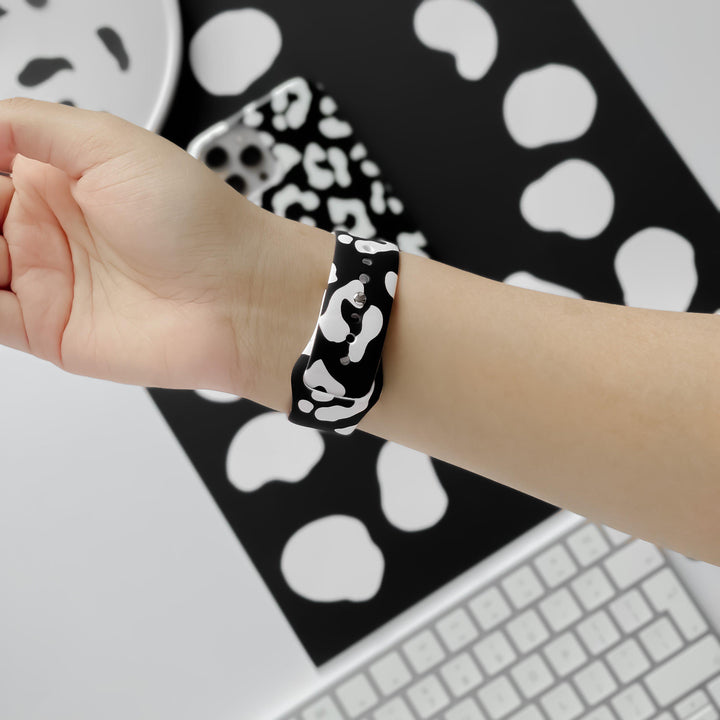 A person’s hand adorned with a wristband from Coconut Lane's Black Leopard Collection is displayed against a backdrop featuring a matching black and white abstract design. A keyboard and phone featuring similar patterns are seen in the background, highlighted by the striking Black Leopard Apple Watch Strap from Coconut Lane.