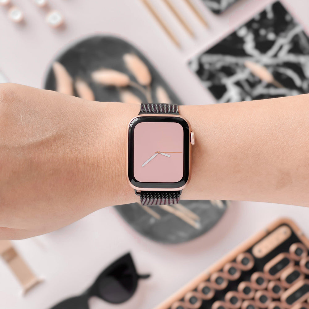 A person wearing an Apple Watch with a pink face and the Coconut Lane Magnetic Chain Apple Watch Strap - Black, featuring a sleek stainless steel clasp. The background showcases various items, including a keyboard, sunglasses, pens, and a patterned notebook, all in coordinated shades of pink, black, and white.