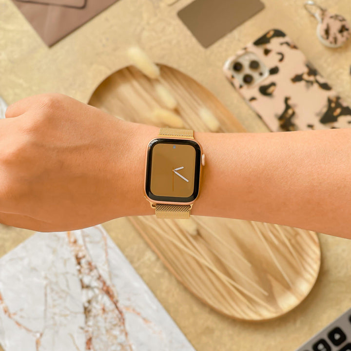 A person wearing a Coconut Lane Magnetic Chain Apple Watch Strap in Gold on their wrist. The watch face displays the time with a simplistic, minimalistic design. In the background, various items such as a phone with a patterned case, wooden tray, and decorative materials are visible.