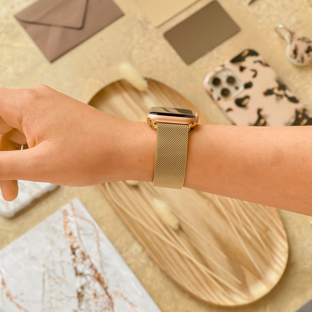 A hand adorned with the Magnetic Chain Apple Watch Strap - Gold by Coconut Lane is extended over a flat lay of items, including a wooden tray, a leopard print phone case, decorative pieces, and various envelopes. The background is warm beige with an array of textures and patterns.