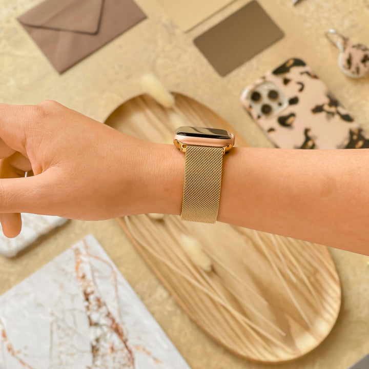 A hand adorned with the Magnetic Chain Apple Watch Strap - Gold by Coconut Lane is extended over a flat lay of items, including a wooden tray, a leopard print phone case, decorative pieces, and various envelopes. The background is warm beige with an array of textures and patterns.