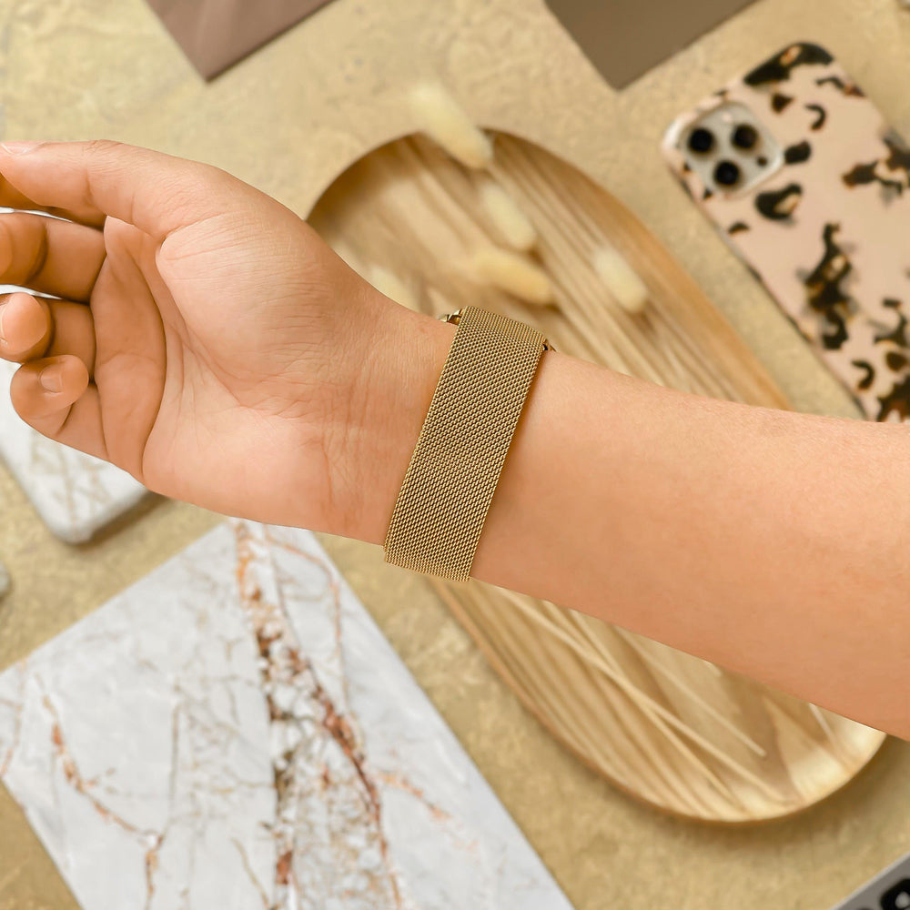 A person's wrist adorned with Coconut Lane's Magnetic Chain Apple Watch Strap in gold, the hand slightly tilted upwards. In the background, a beige phone case with a brown and black pattern, rectangular plates featuring white marble designs, and an oblong wooden tray with decorative items are visible.