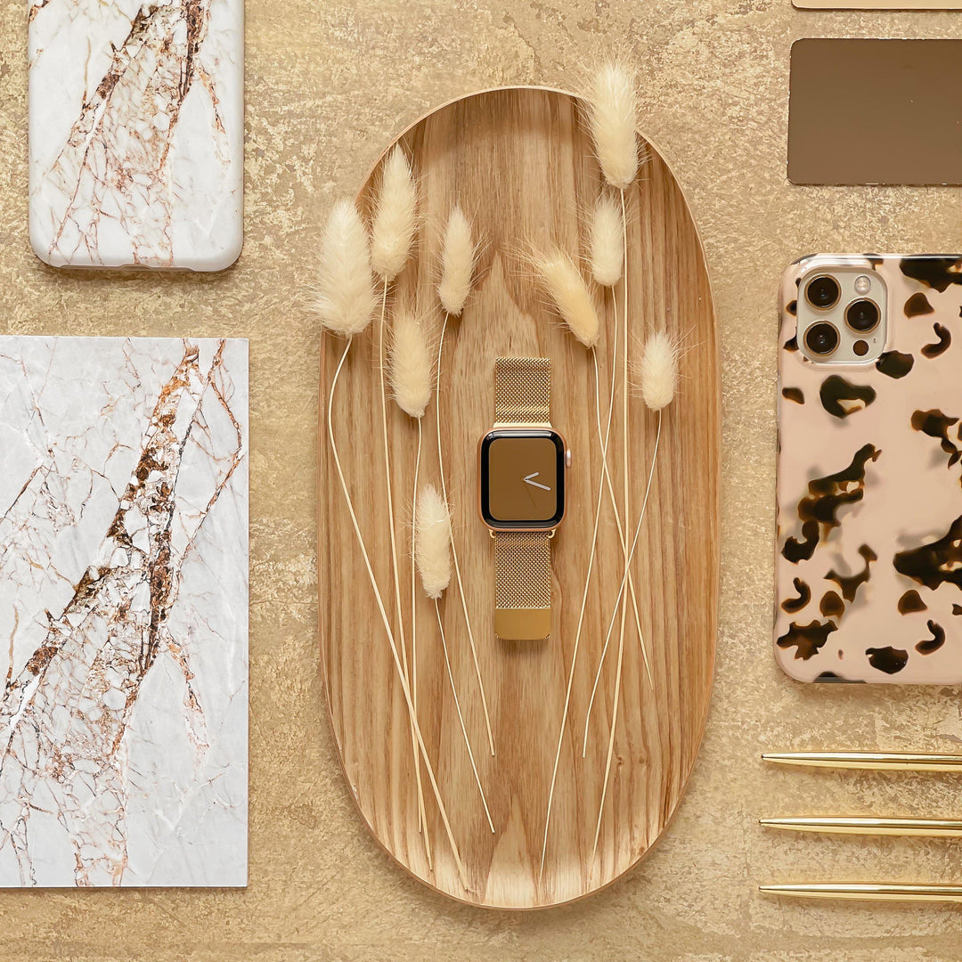 A stylish flat lay features the Coconut Lane Magnetic Chain Apple Watch Strap in gold with a stainless steel magnetic clasp, elegantly placed on a wooden tray adorned with decorative dried grasses. Surrounding items include two marble-patterned notebooks, a leopard print phone case, and gold stationery set against a beige textured background.
