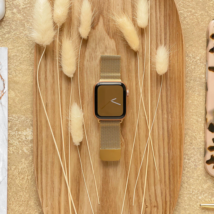 A gold Coconut Lane Magnetic Chain Apple Watch Strap is placed on an oval wooden tray, surrounded by dried beige fluffy grasses. The background features a soft golden-textured surface and parts of other items, including a marble-patterned object and a tortoiseshell object.