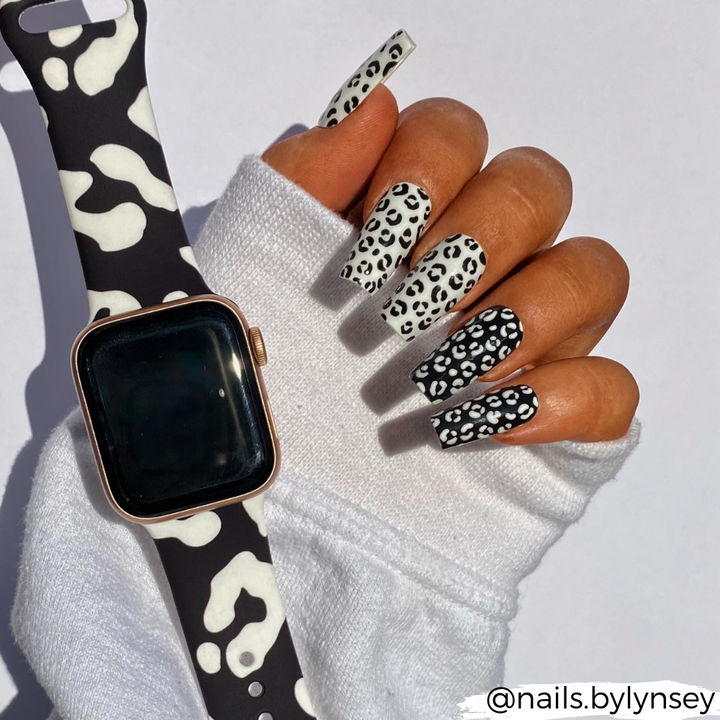 A hand with long square-shaped nails showcases a black and white cow print manicure. The individual is adorned in a white long sleeve shirt and sports a Black Leopard Apple Watch Strap by Coconut Lane. The Instagram handle "@nails.bylynsey" is visible at the bottom right corner.