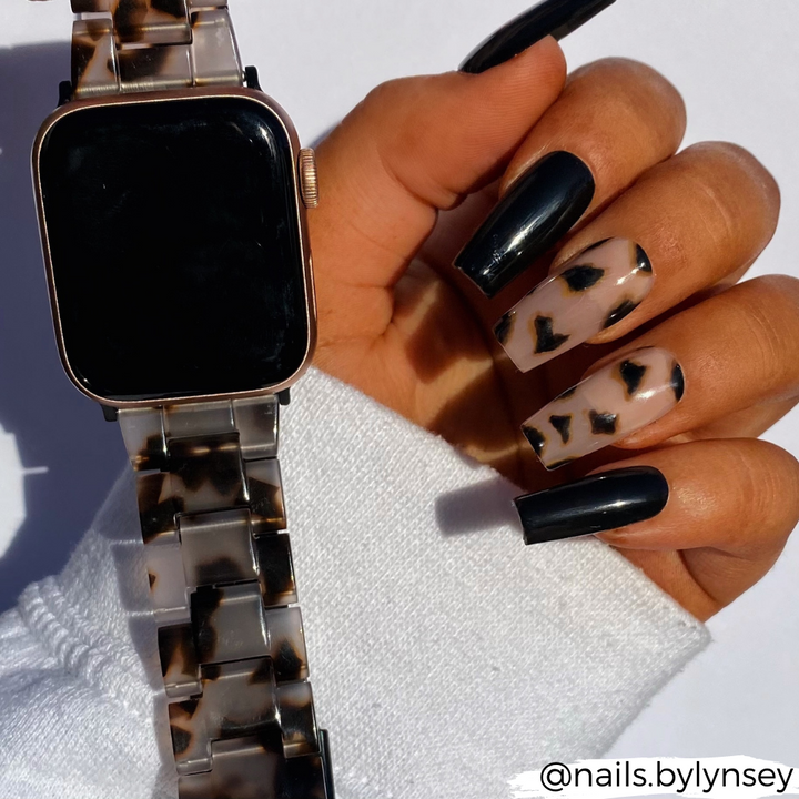 A hand with nails painted in a black and tortoiseshell pattern holds an arm up, showcasing the Luxe Ivory Tort Apple Watch Strap by Coconut Lane, designed with a matching tortoiseshell pattern. The person is dressed in a white long-sleeve top. The Instagram handle @nails.bylynsey is visible in the bottom right corner.