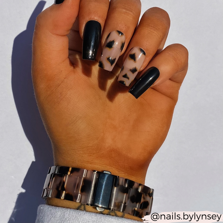 A hand with long, square-shaped nails featuring a manicure is adorned with solid black polish, accented by two nails with a clear base and black abstract spots. The person is wearing the Luxe Ivory Tort Apple Watch Strap by Coconut Lane. The background is white.
