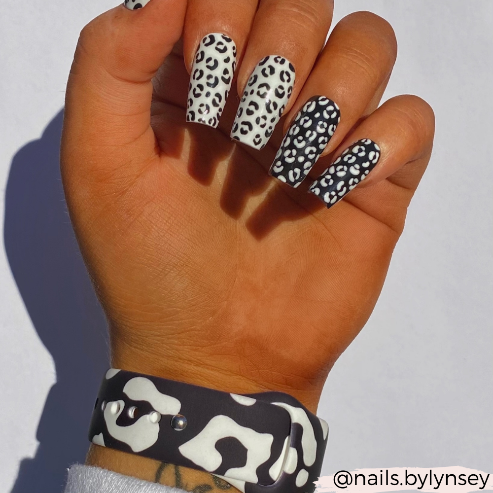 A hand with long, square-shaped nails showcases a black and white leopard print design. The nails alternate between white with black spots and black with white spots. The wrist elegantly displays the stylish Black Leopard Apple Watch Strap by Coconut Lane. The background is a pure, clean white.
