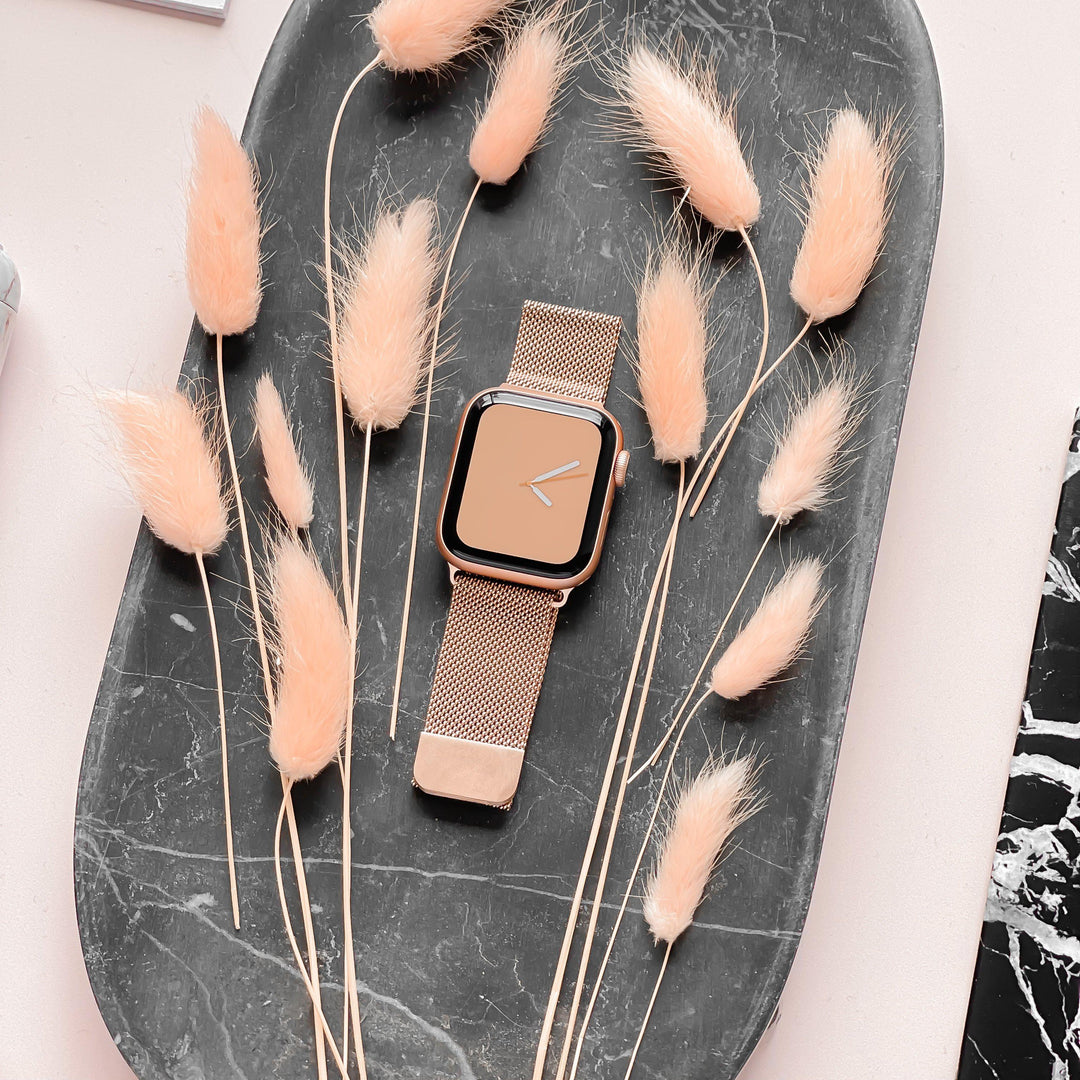 A rectangular Apple Watch model with a Magnetic Chain Apple Watch Strap in Rose Gold from Coconut Lane is placed on an oval dark gray tray along with beige dried grasses, artfully arranged around it. The background is pale pink.