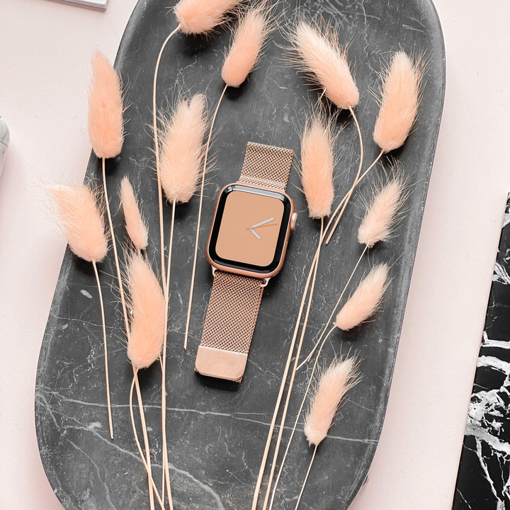 A rectangular Apple Watch model with a Magnetic Chain Apple Watch Strap in Rose Gold from Coconut Lane is placed on an oval dark gray tray along with beige dried grasses, artfully arranged around it. The background is pale pink.