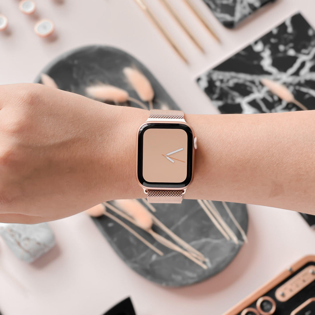 A person wearing an Apple Watch adorned with a minimalist clock face and the Magnetic Chain Apple Watch Strap - Rose Gold from Coconut Lane on their wrist. The background features various aesthetically arranged objects such as a marbled tray, plants, and light-colored items.