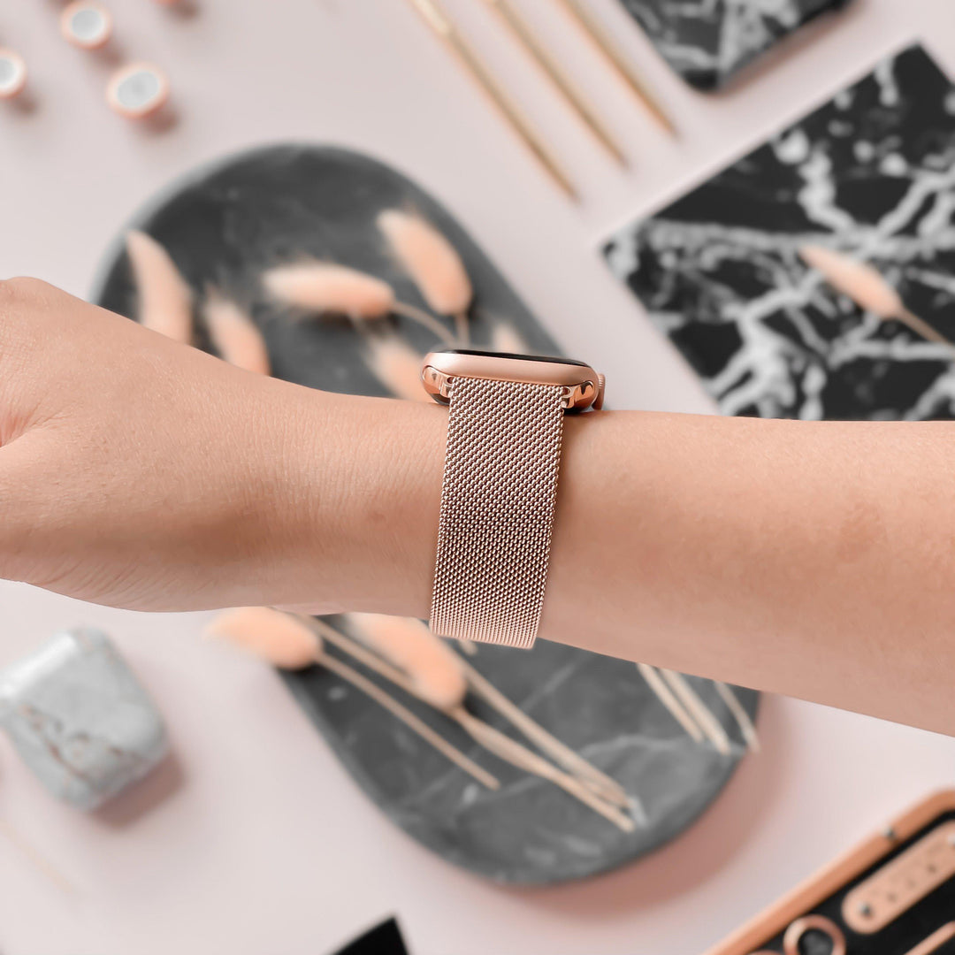 A person wearing a Coconut Lane Magnetic Chain Apple Watch Strap in Rose Gold, viewed from the back and set against a stylish background with marble-patterned items, small decor pieces, and stationery. The arm and wrist are the main focus of the image, highlighting the elegance akin to Rose Gold Magnetic Chain accessories.
