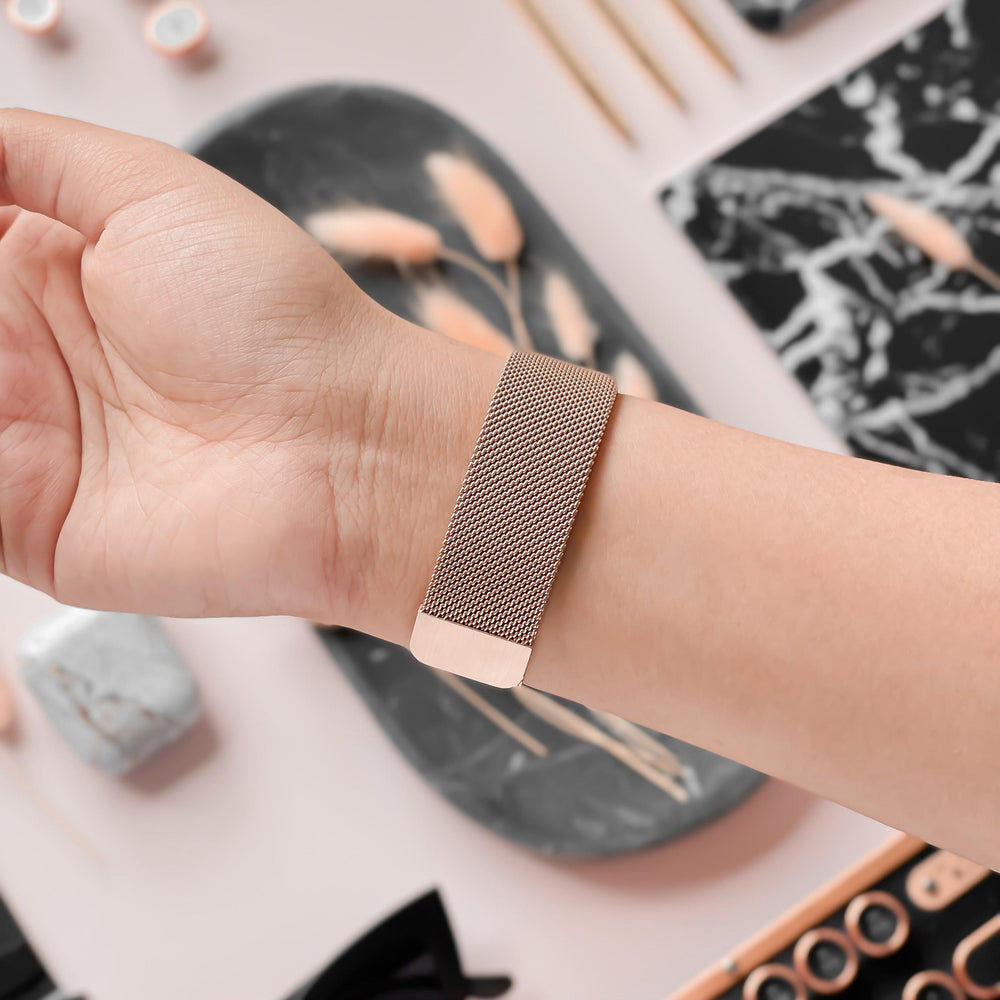 A close-up of a person's wrist wearing Coconut Lane's stylish Magnetic Chain Apple Watch Strap in Rose Gold. The background features a marble tray adorned with dried flowers, wooden sticks, and various small desk items like scissors and paper clips, all arranged neatly with an aesthetically pleasing setup.
