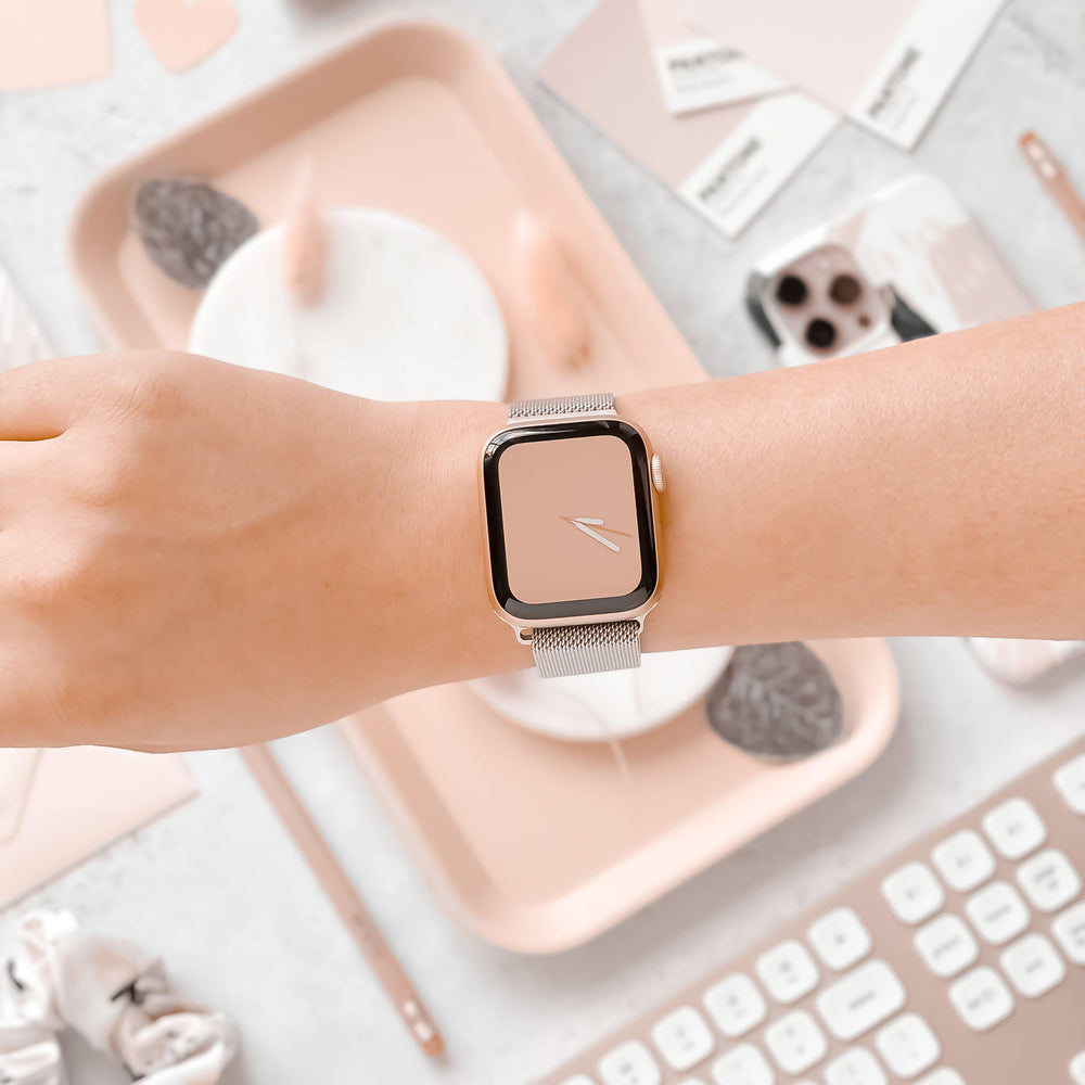 A close-up of a person's wrist adorned with the Coconut Lane Magnetic Chain Apple Watch Strap in silver, featuring a modern, rectangular smartwatch with a pink face and a sleek silver mesh band that has a Stainless Steel Magnetic Clasp. The background highlights a pastel-themed workspace filled with various stationery, a computer keyboard, and décor items in soft pink and gray tones.