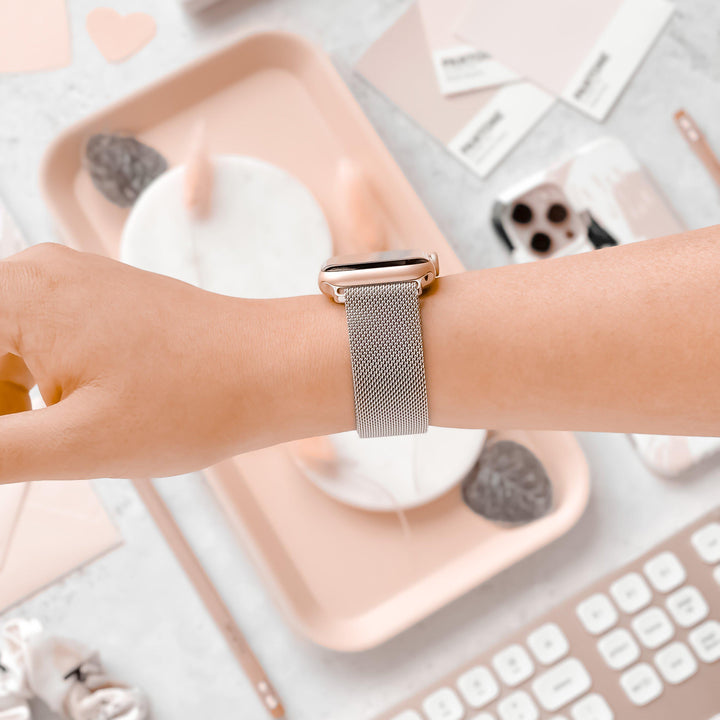 A hand wearing Coconut Lane’s stylish Magnetic Chain Apple Watch Strap in silver with a sleek stainless steel magnetic clasp is seen against a backdrop of neatly arranged office items. The background includes a keyboard, some documents, a tray with rocks and candles, and a palette of pink and neutral colors, creating a visually pleasing scene.