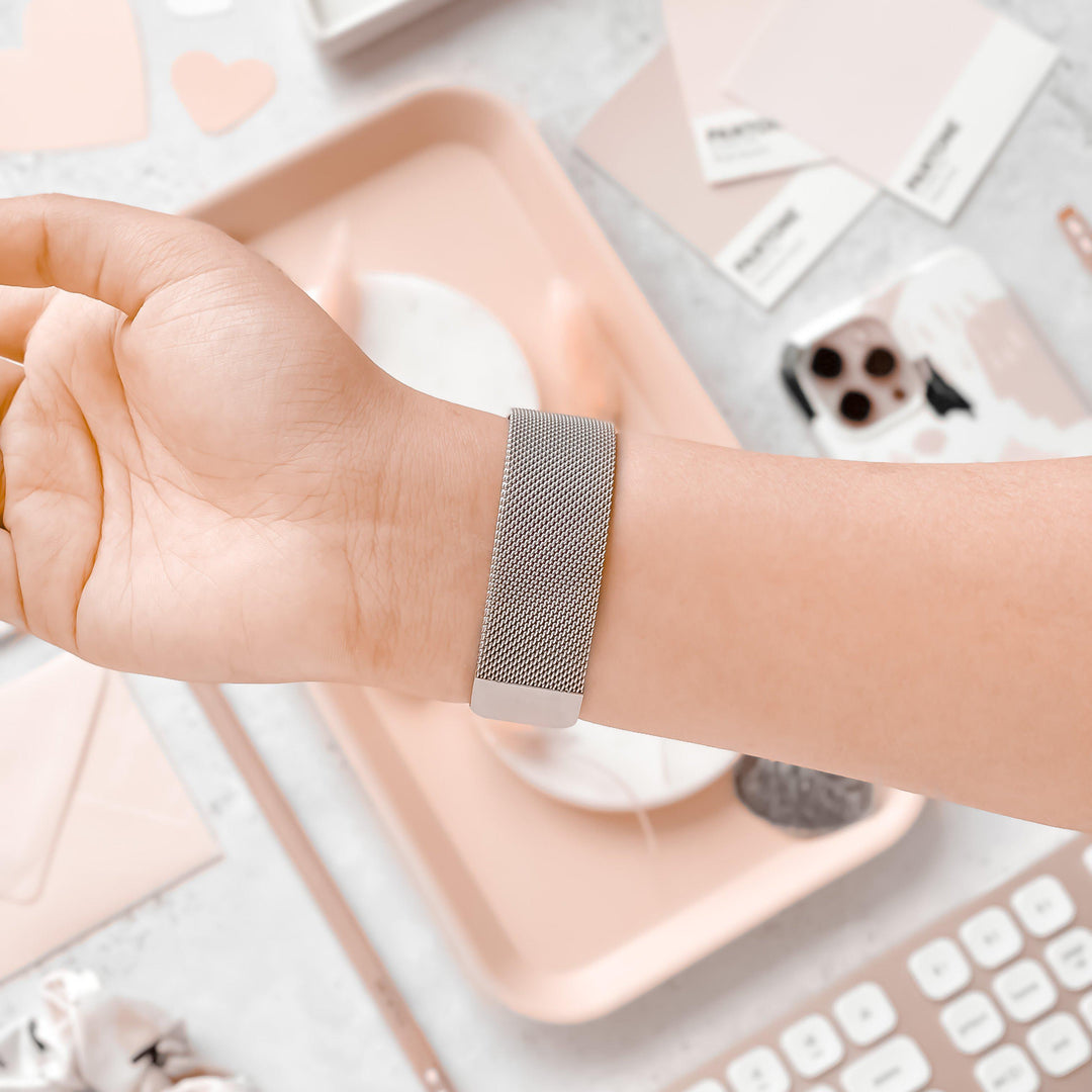 A person wearing a Coconut Lane Magnetic Chain Apple Watch Strap in Silver on their wrist. The background features a workspace with items in shades of pink, including a tray, paint swatches, paper hearts, and a keyboard. The scene has a soft, aesthetically pleasing ambiance.